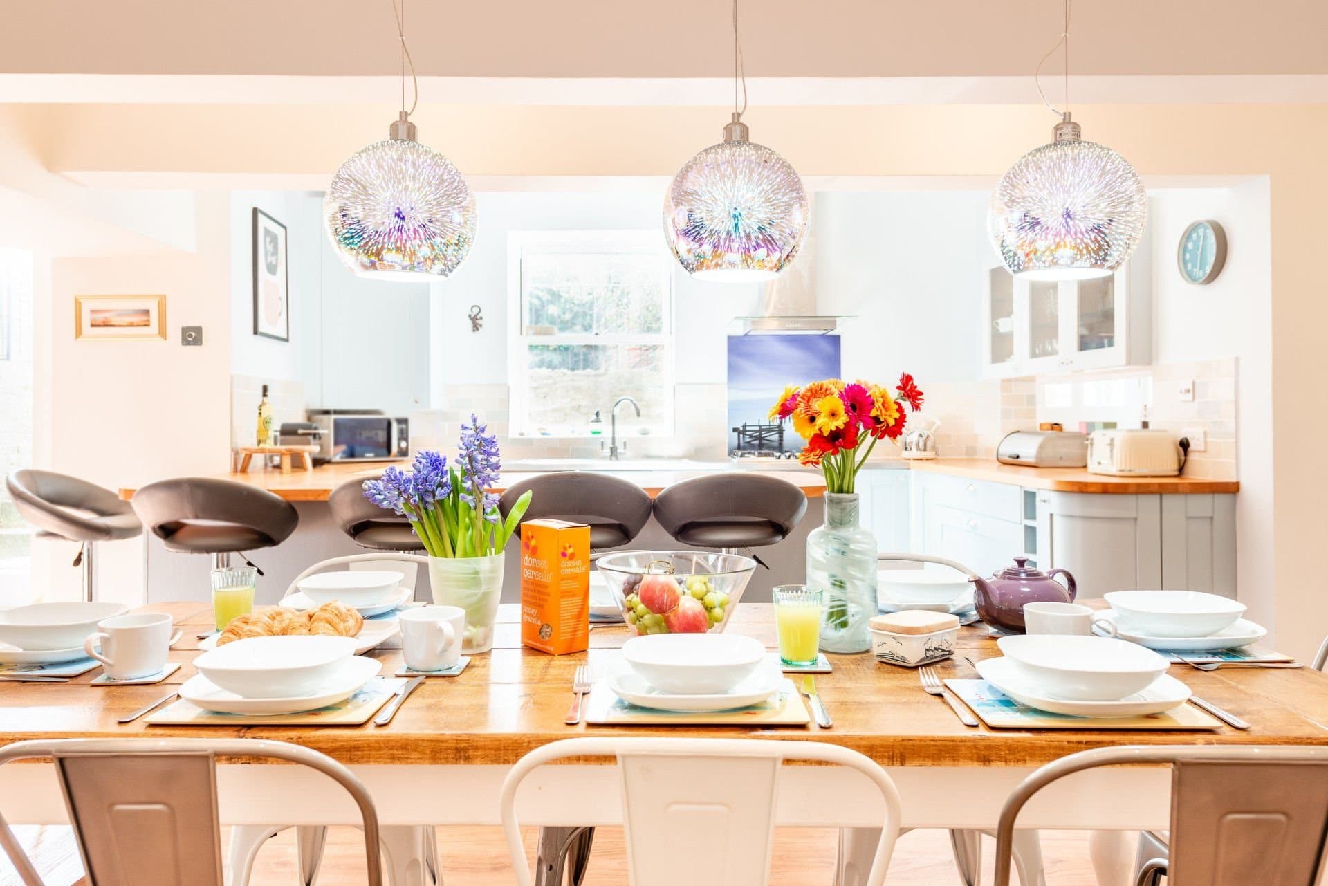 Dining table set for breakfast, with the kitchen and iridescent pendant lights beyond